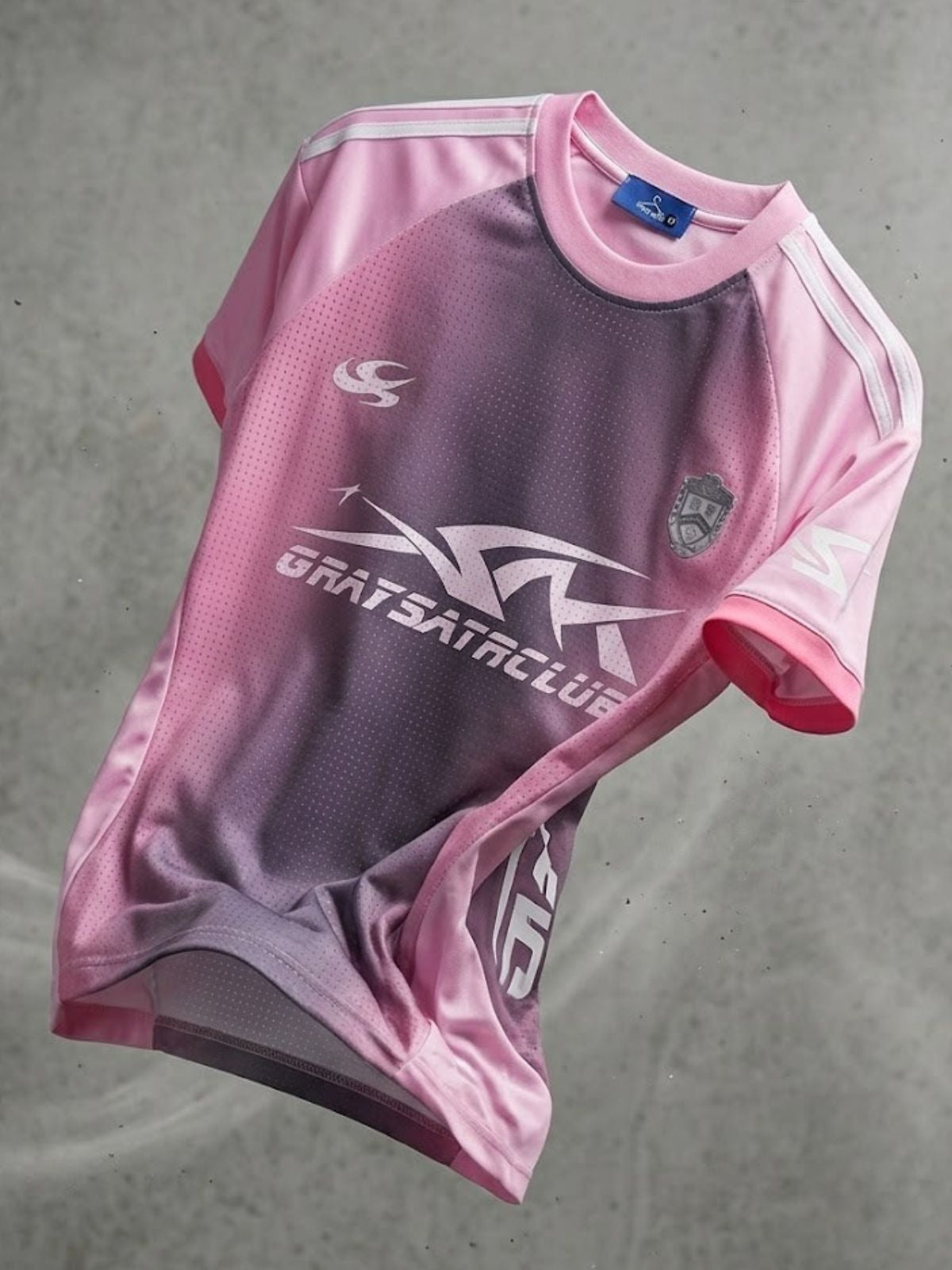 A pink and purple gradient athletic jersey with white graphics, including the text 'GRATSACLUDE' and a club crest, laid flat on a gray concrete surface.